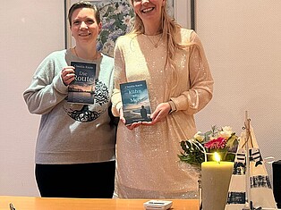 Die Autorin Frau Raute (links) und Frau Baumann von der Kulturarbeit (rechts) stehen hinter dem Vorlesetisch und halten jeweils ein Buch hoch. Einmal "Die Route" und einmal "Kühn am Meer", beide von Frau Raute geschrieben.