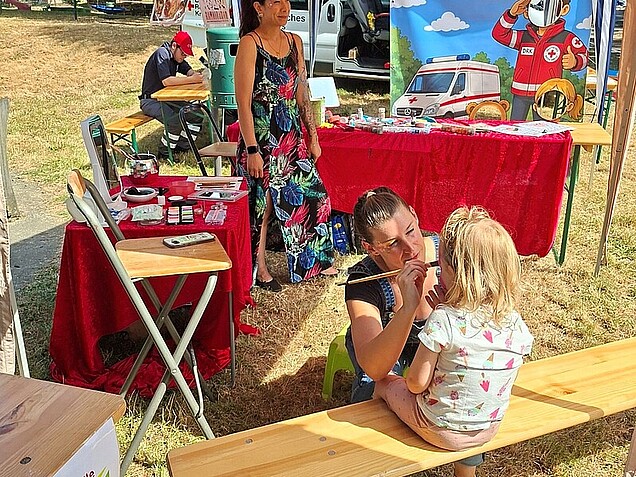 An der Stempelstelle an der St. Johann Promenade in Bergshausen wird ein Kind geschminkt. Im Hintergrund ist ein Schminkstand, sowie ein Infostand vom DRK zu sehen.