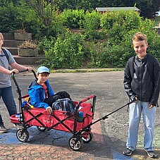 Foto von zwei Kindern die einen Bollerwagen schieben. Im Bollerwagen sitzt ein weiteres Kind
