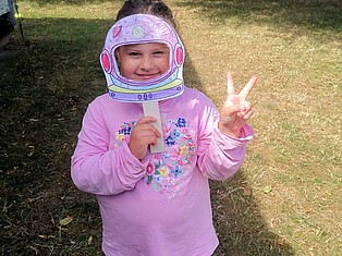 Ein kleines Mädchen steht auf der Wiese und hält sich ihren selbstgebastelten Astronautenhelm vor das Gesicht.