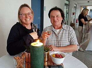 Besucherinnen stehen an einem mit Blumen und Kerzen dekorierten Stehtisch mit einem Sektglas in der Hand.