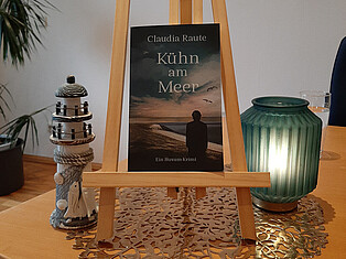 Das Buch der Lesung "Kühn am Meer" von Claudia Raute auf einer kleinen Staffelei auf dem Vorlesetisch. Links davon steht ein kleiner Leuchtturm zur Dekoration und rechts ein Teelicht in einem grünen Glas.