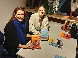 Zwei Damen sitzend am Tisch. Sie präsentieren die Bücher Sunny-Saga "Gebrochene Herzen" und Sunny-Saga "Verschlungene Wege"