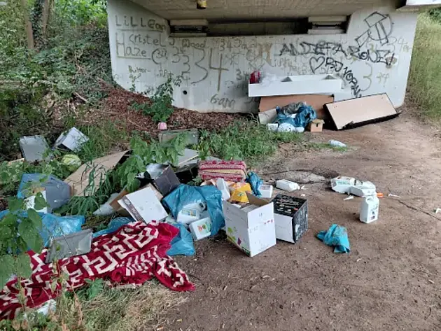 Unter einer Brücke liegt vor einem breiten Brückenpfeiler illegal abgeladener Müll, unter anderem Pappkartons, Plastikmüllsäcke und Möbelteile. 