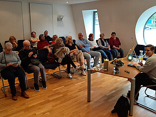 Frau Raute sitzt am Vorlesetisch, ihr gegenüber sitzen 13 Besucherinnen und Besucher. Blick aus Richtung Frau Raute