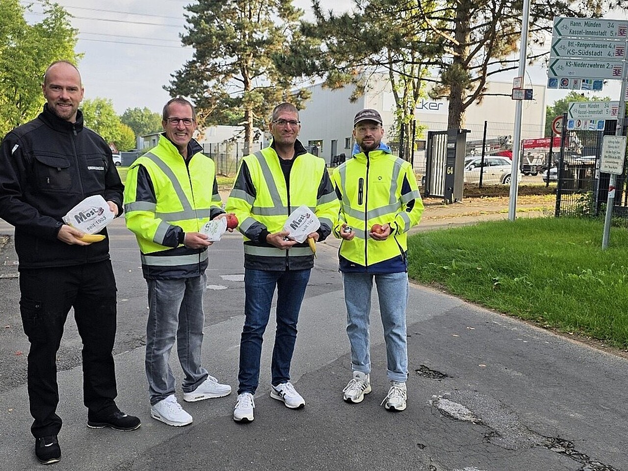 Das Team der Aktion "Pendlerfrühstück": v.l. Dennis Schiffer (Ordnungspolizei); Andreas Damm (Bürgermeister); Oliver Brandenstein (Fachbereichsleiter 4 Bürgerdienstleistungen); Jonas Döbel (Klimaschutzmanager Gemeinde Fuldabrück) stehen in Warnjacken an der Straße und halten Brezeln und Äpfel in der Hand
