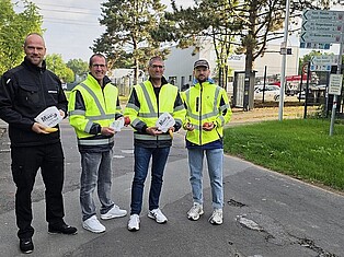Das Team der Aktion "Pendlerfrühstück": v.l. Dennis Schiffer (Ordnungspolizei); Andreas Damm (Bürgermeister); Oliver Brandenstein (Fachbereichsleiter 4 Bürgerdienstleistungen); Jonas Döbel (Klimaschutzmanager Gemeinde Fuldabrück) stehen in Warnjacken an der Straße und halten Brezeln und Äpfel in der Hand